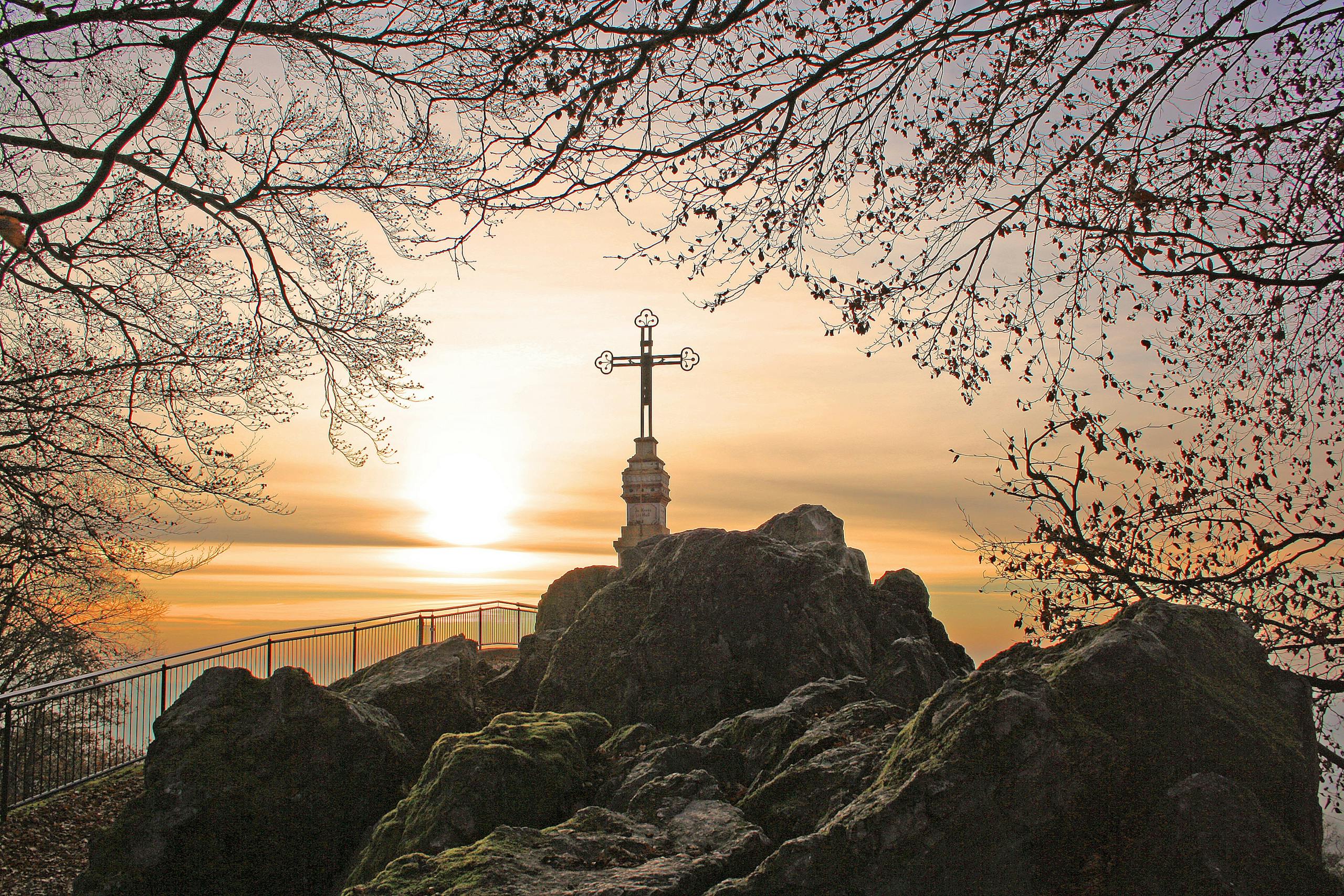 A captivating sunrise view with a cross atop a rocky summit, surrounded by trees.